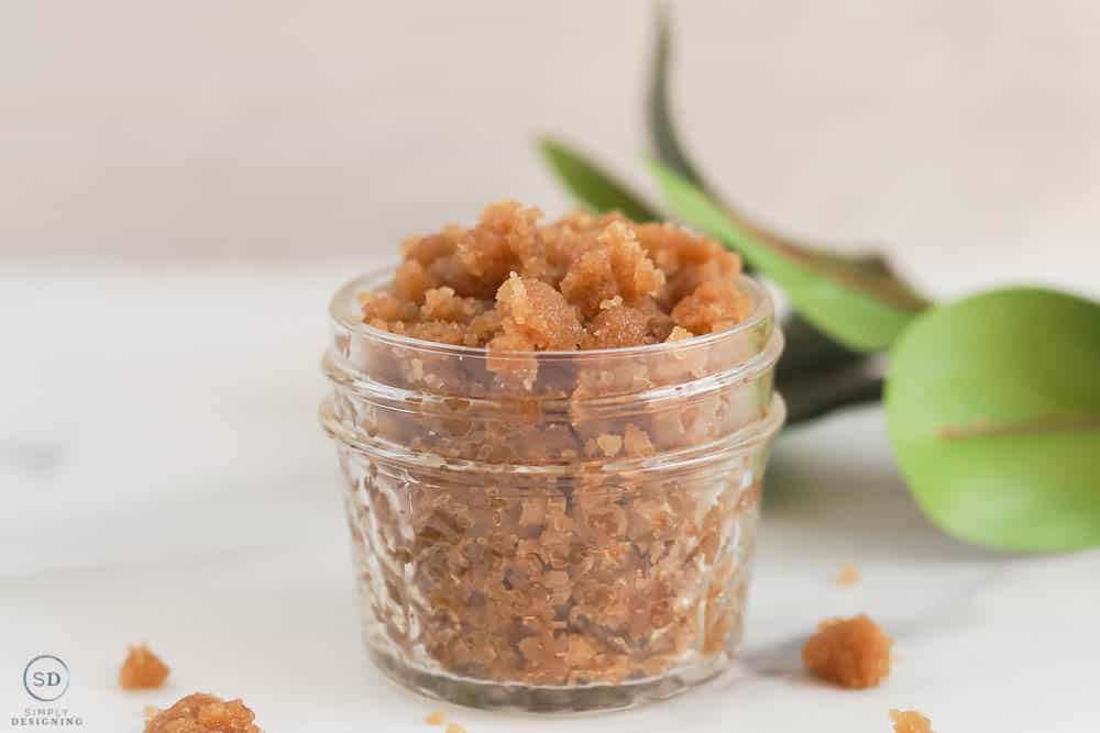 homemade sugar scrub with brown sugar in a glass jar overflowing looking at it from the side in a horizontal photo with greenery in the background